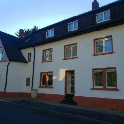 Dwukondygnacyjny budynek z białymi ścianami i ciemnoczerwoną lamperią oraz obramowaniami okien, sfotografowany z poziomu ulicy w słoneczny dzień.