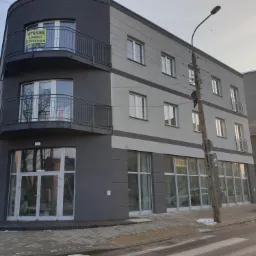 Nowoczesny budynek biurowy z balkonami, pomalowany w odcieniach szarości, z widocznymi oknami i drzwiami wejściowymi, na jednym z balkon&oacute;w wywieszona tablica informująca o wynajmie lokali użytkowych.