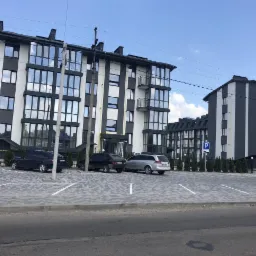 Nowoczesny budynek wielorodzinny w Lublinie z parkingiem wyłożonym kostką brukową, utrzymany w tonacji bieli i szarości, z licznymi oknami i balkonami.