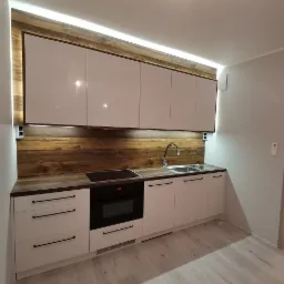 Nowoczesna kuchnia z białymi, lakierowanymi frontami, drewnianym panelem nad blatem i podświetleniem LED, czarne, podłużne uchwyty, płyta indukcyjna i piekarnik w zabudowie.