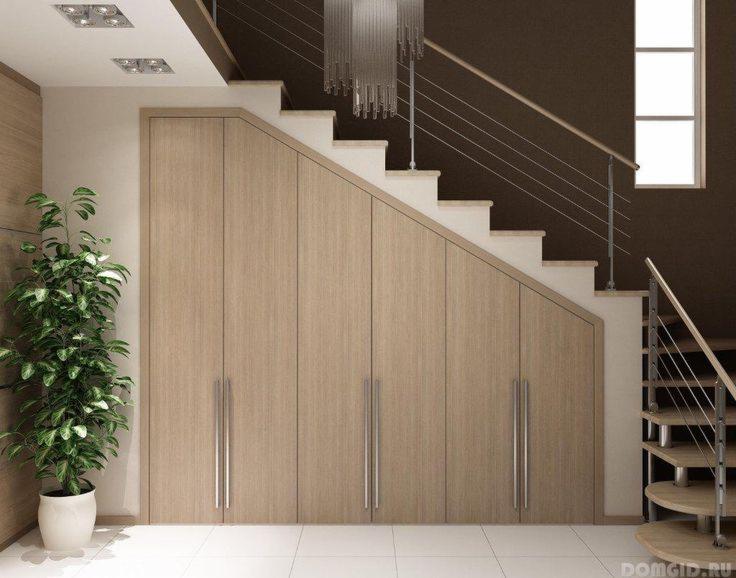 Drewniana szafa wnękowa pod schodami z czterema drzwiami i podłużnymi uchwytami, obok doniczka z zieloną rośliną, spiralne schody z metalową poręczą i wiszący żyrandol.