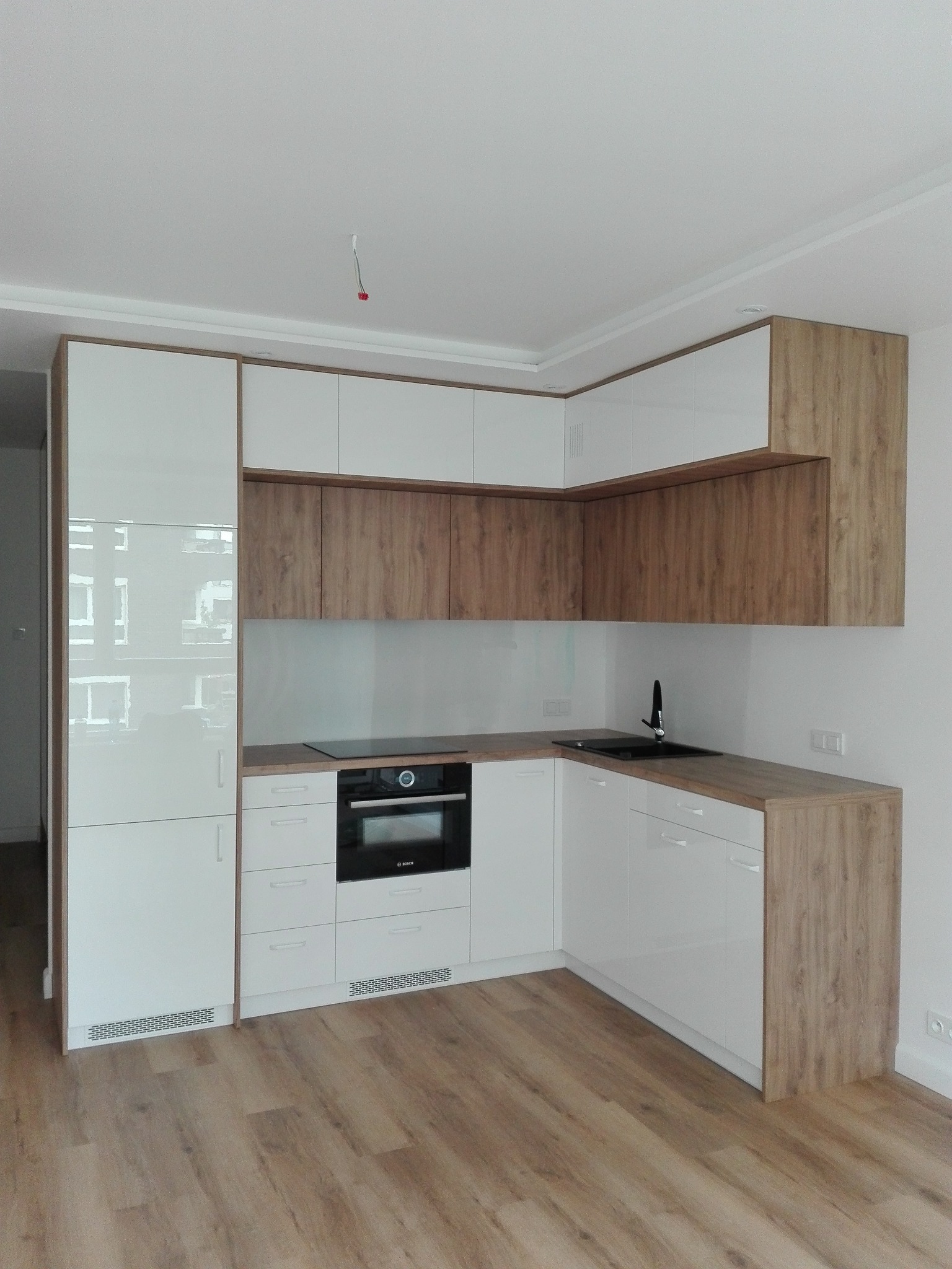 Nowoczesna, narożna kuchnia z białymi, lakierowanymi frontami i okładzinami w kolorze dębu amerykańskiego, wyposażona w płytę indukcyjną i piekarnik marki Bosch.