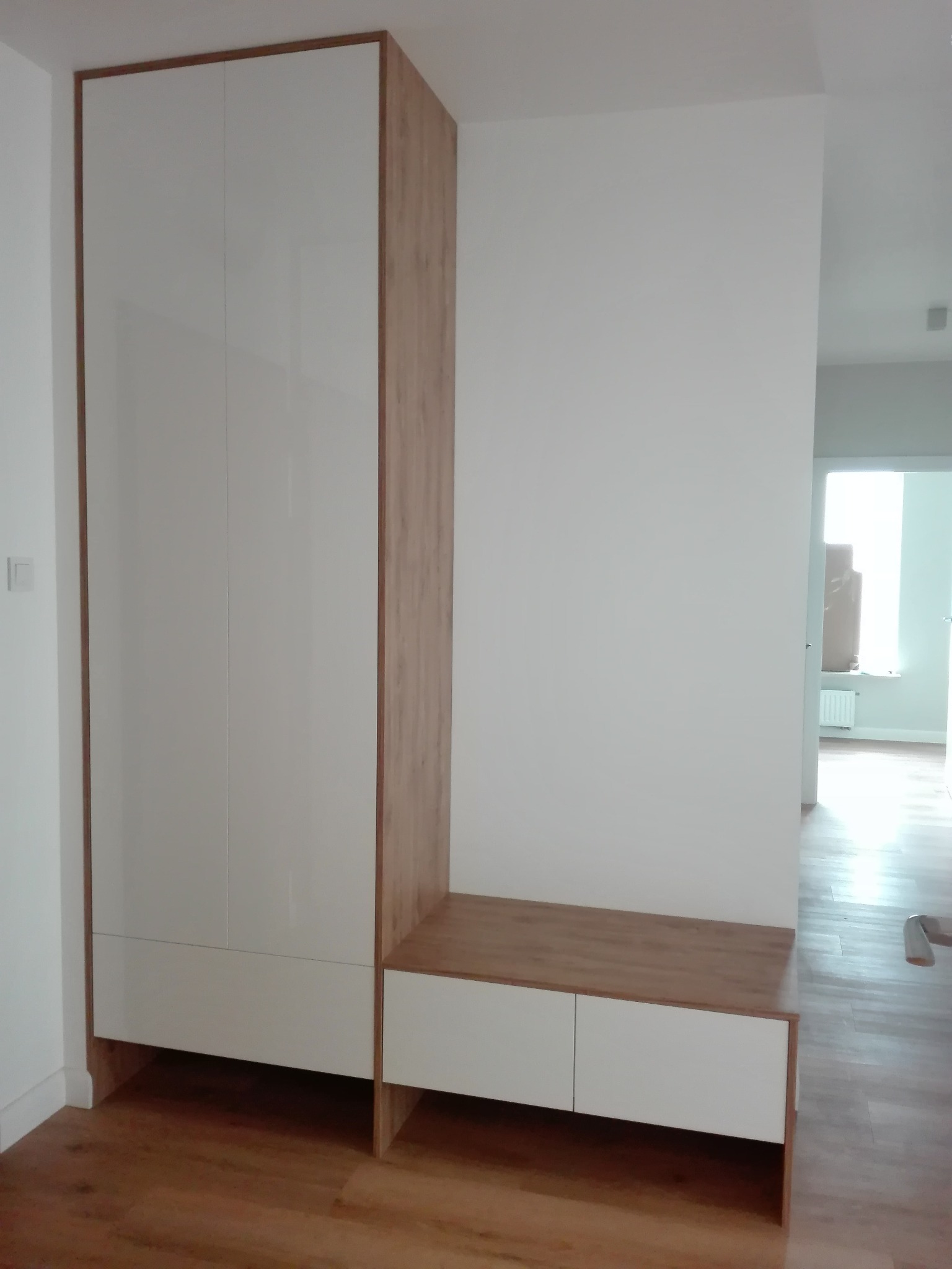 Szafa w przedpokoju z białymi, lakierowanymi frontami na wysoki połysk, drewnopodobnym korpusem i wbudowaną niską szafką z siedziskiem, z widoczną częścią podłogi z paneli.