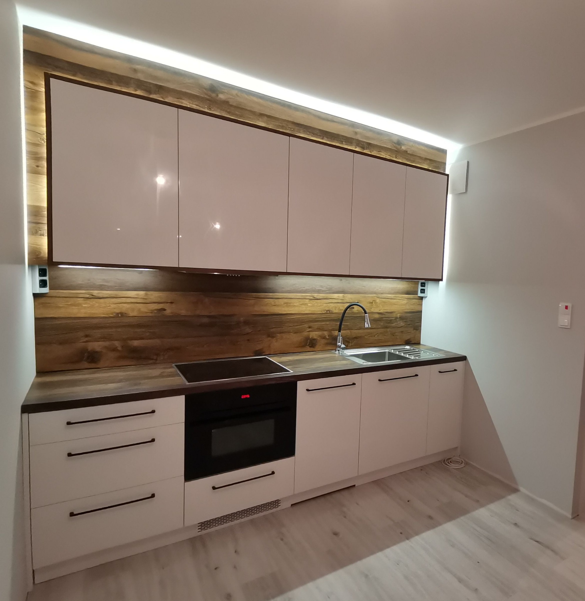 Nowoczesna kuchnia z białymi, lakierowanymi frontami, drewnianym panelem nad blatem i podświetleniem LED, czarne, podłużne uchwyty, płyta indukcyjna i piekarnik w zabudowie.