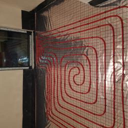 Bohdan - Czerwone rury ogrzewania podłogowego przymocowane do siatki na ścianie, widoczne okno w lewym górnym rogu.