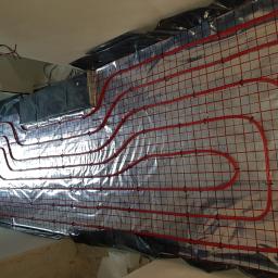 Bohdan - Czerwone rury systemu ogrzewania podłogowego ułożone na srebrnej folii izolacyjnej z czerwoną siatką, zabezpieczone czarną folią przy ścianach w surowym wnętrzu.