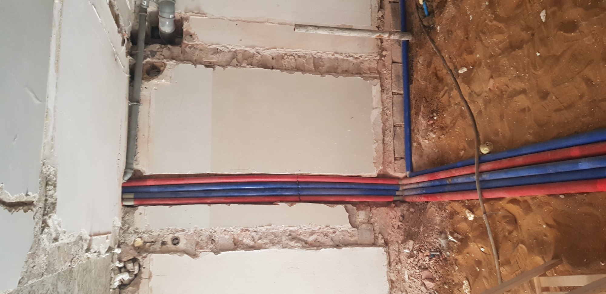 Wykop pod podłogą w trakcie prac remontowych w Warszawie, widoczne rury instalacji wodnej i elektrycznej w bruzdach w ścianie, częściowo zakryte tynkiem.