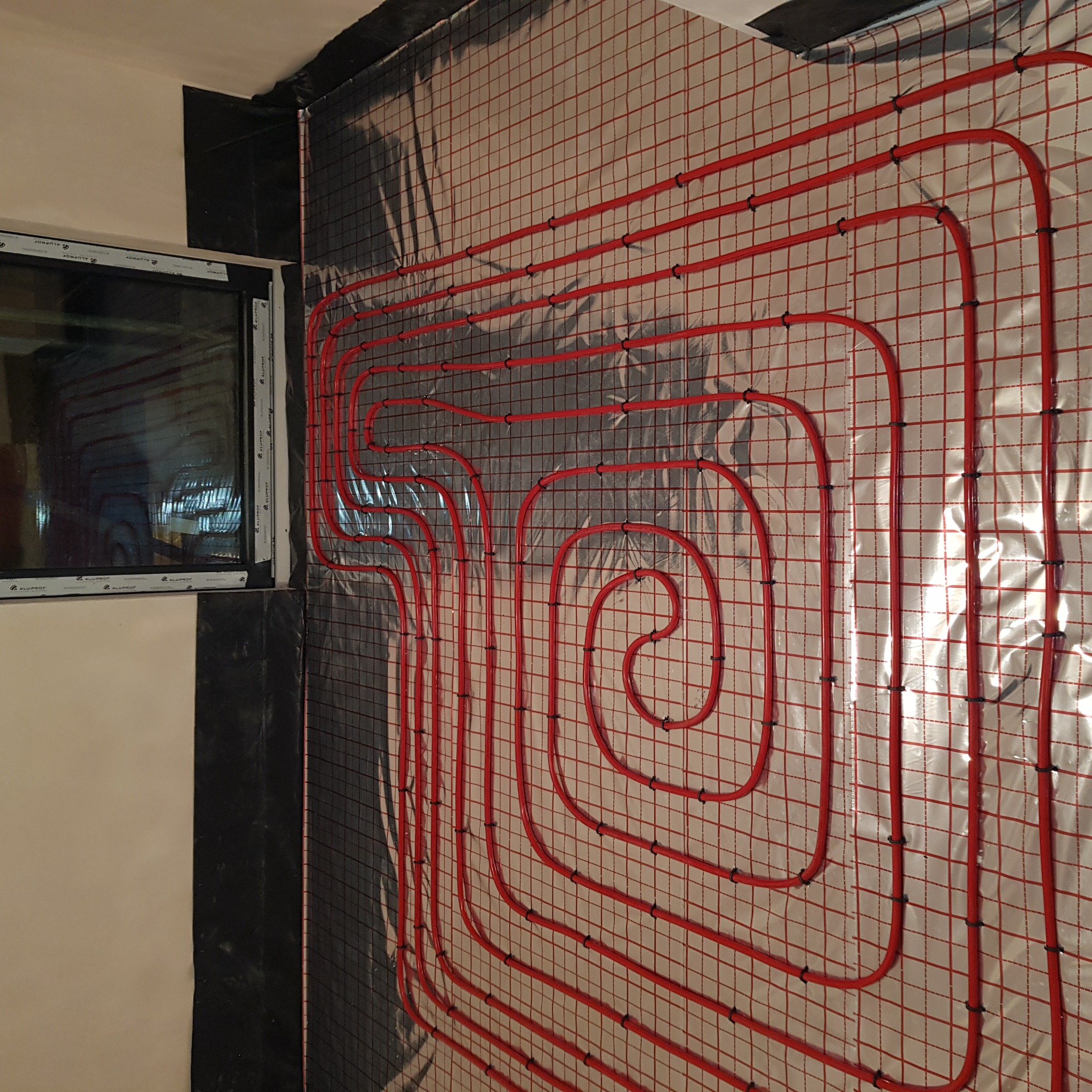 Czerwone rury ogrzewania podłogowego przymocowane do siatki na ścianie, widoczne okno w lewym górnym rogu.