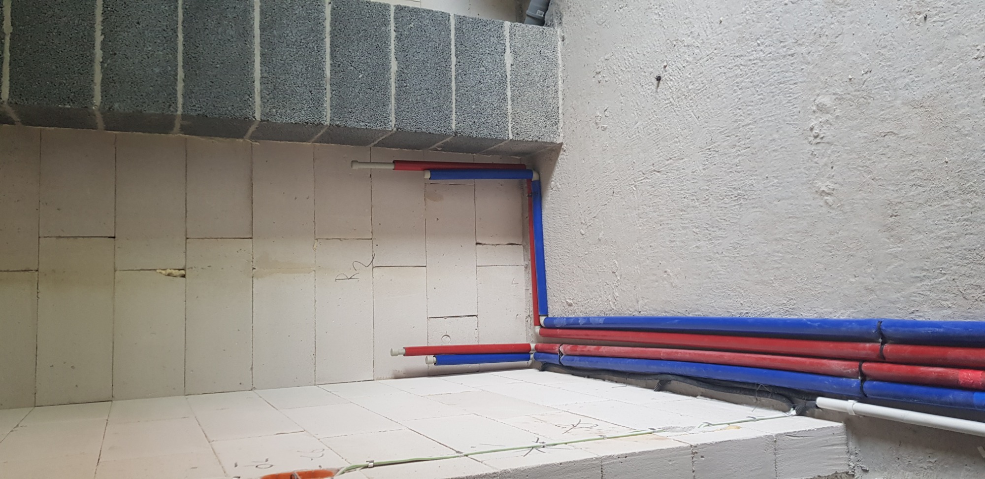 Fragment ściany z widocznymi przewodami elektrycznymi w kolorze czerwonym i niebieskim, ułożonymi wzdłuż muru z białych bloczków.