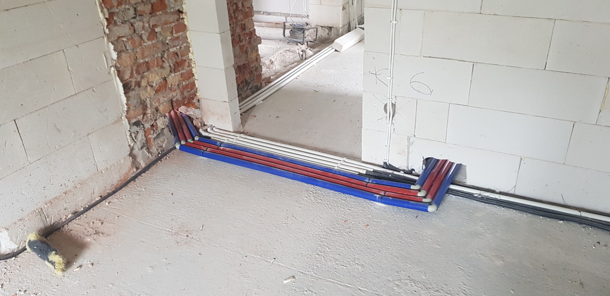 Wiązka przewodów elektrycznych w peszlach, prowadzona wzdłuż ściany z białych bloczków, z widocznymi otworami w ścianie i fragmentami instalacji w sąsiednim pomieszczeniu, w surowym stanie.