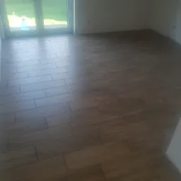 Ułożona podłoga z płytek imitujących drewno w pustym pokoju, widoczne narzędzia i butelka w rogu.