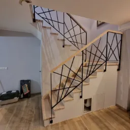 Nowoczesne schody wewnętrzne z drewnianymi stopniami i czarną, geometryczną balustradą, widoczne z perspektywy dolnej części biegu, z elementami wykończenia wnętrza w tle.