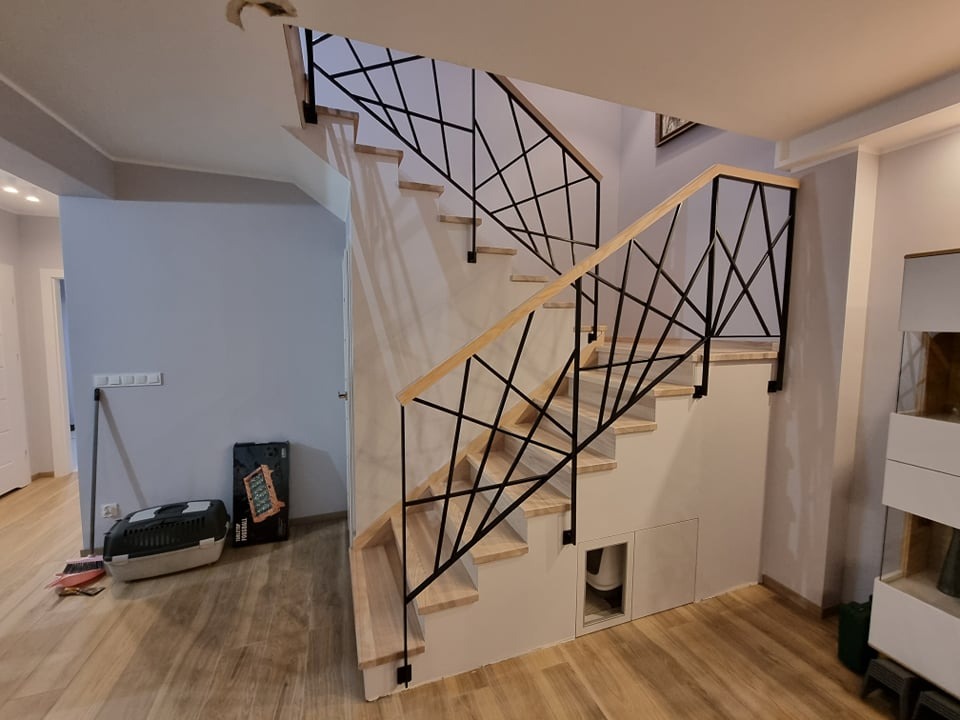 Nowoczesne schody wewnętrzne z drewnianymi stopniami i czarną, geometryczną balustradą, widoczne z perspektywy dolnej części biegu, z elementami wykończenia wnętrza w tle.