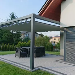 Nowoczesna pergola z rozsuwanym zadaszeniem materiałowym w kolorze szarym, przylegająca do budynku z roletą zewnętrzną i drewnianą okładziną, na wybrukowanym tarasie z kompletem mebli ogrodowych.
