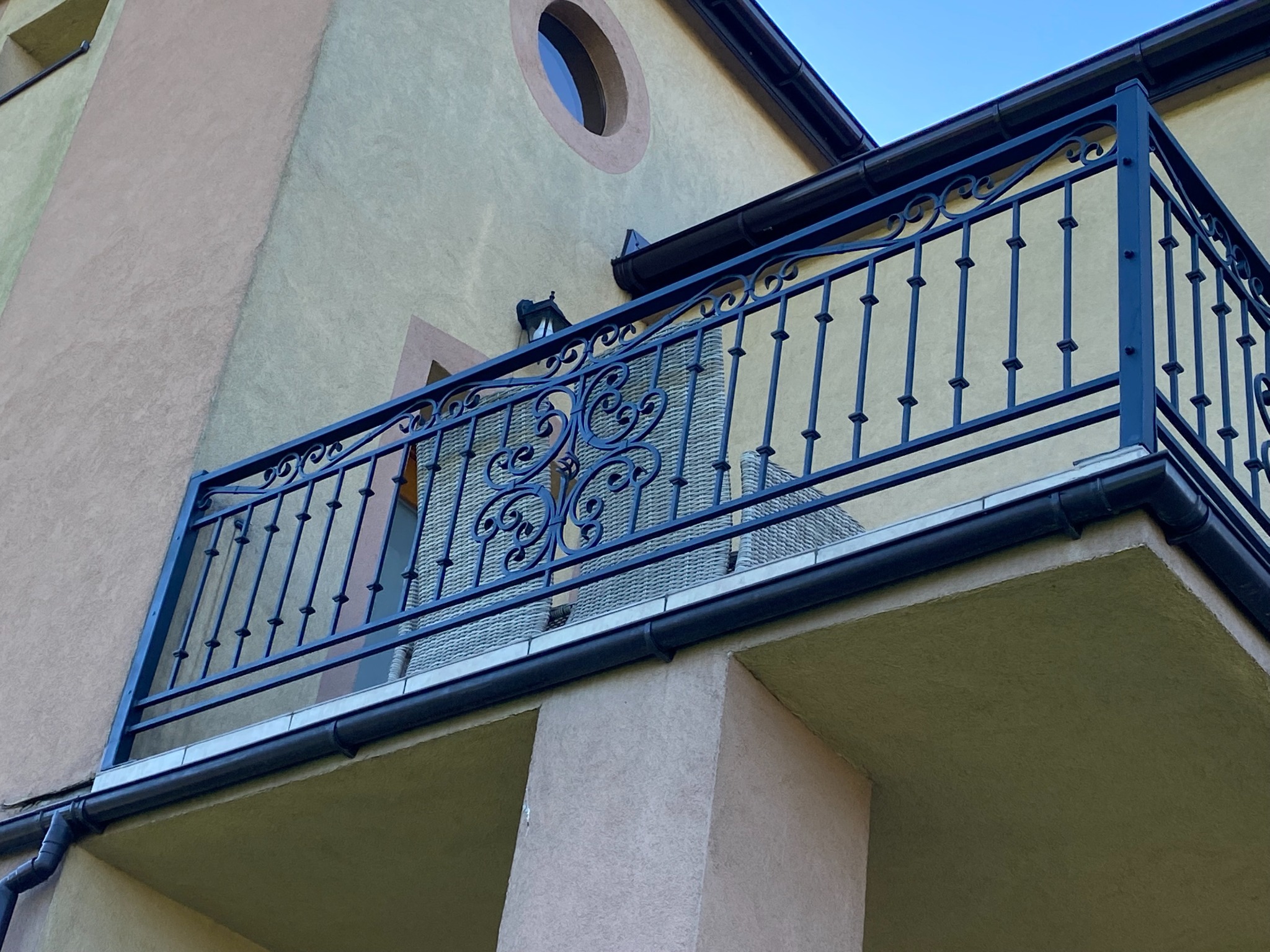 Kuta, czarna balustrada balkonowa z dekoracyjnymi elementami na tle beżowej elewacji budynku z okrągłym oknem.