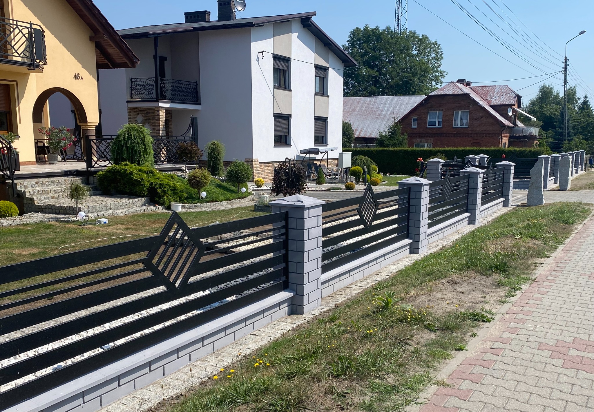 Nowoczesne ogrodzenie z czarnych, poziomych profili z dekoracyjnym rombem i szarymi słupkami z cegieł, oddzielające posesję od chodnika z kostki brukowej.