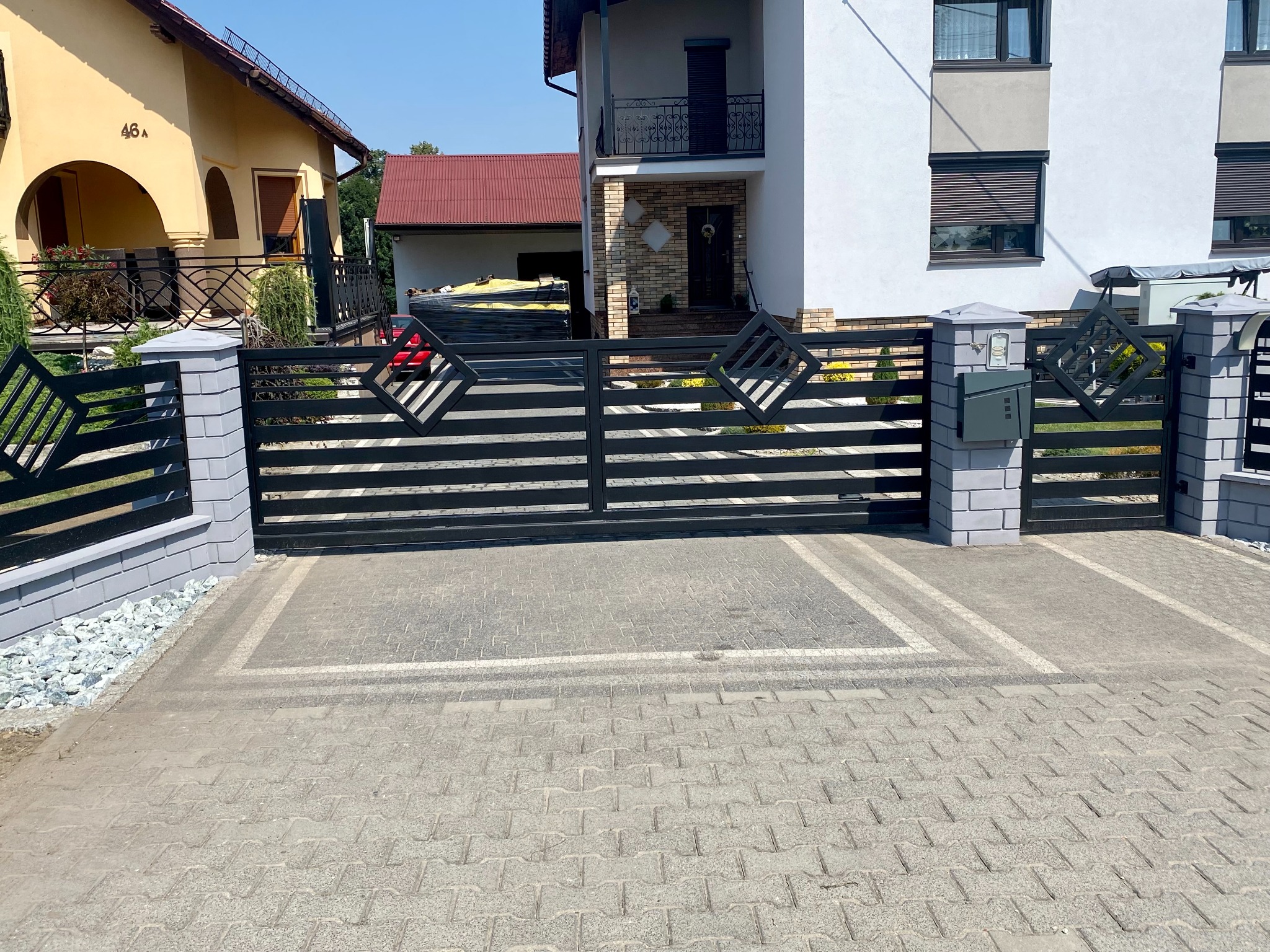 Nowoczesna, przesuwna brama wjazdowa z poziomymi, czarnymi profilami i dekoracyjnymi elementami w kształcie rombów, zamontowana przed domem z szarą skrzynką na listy.