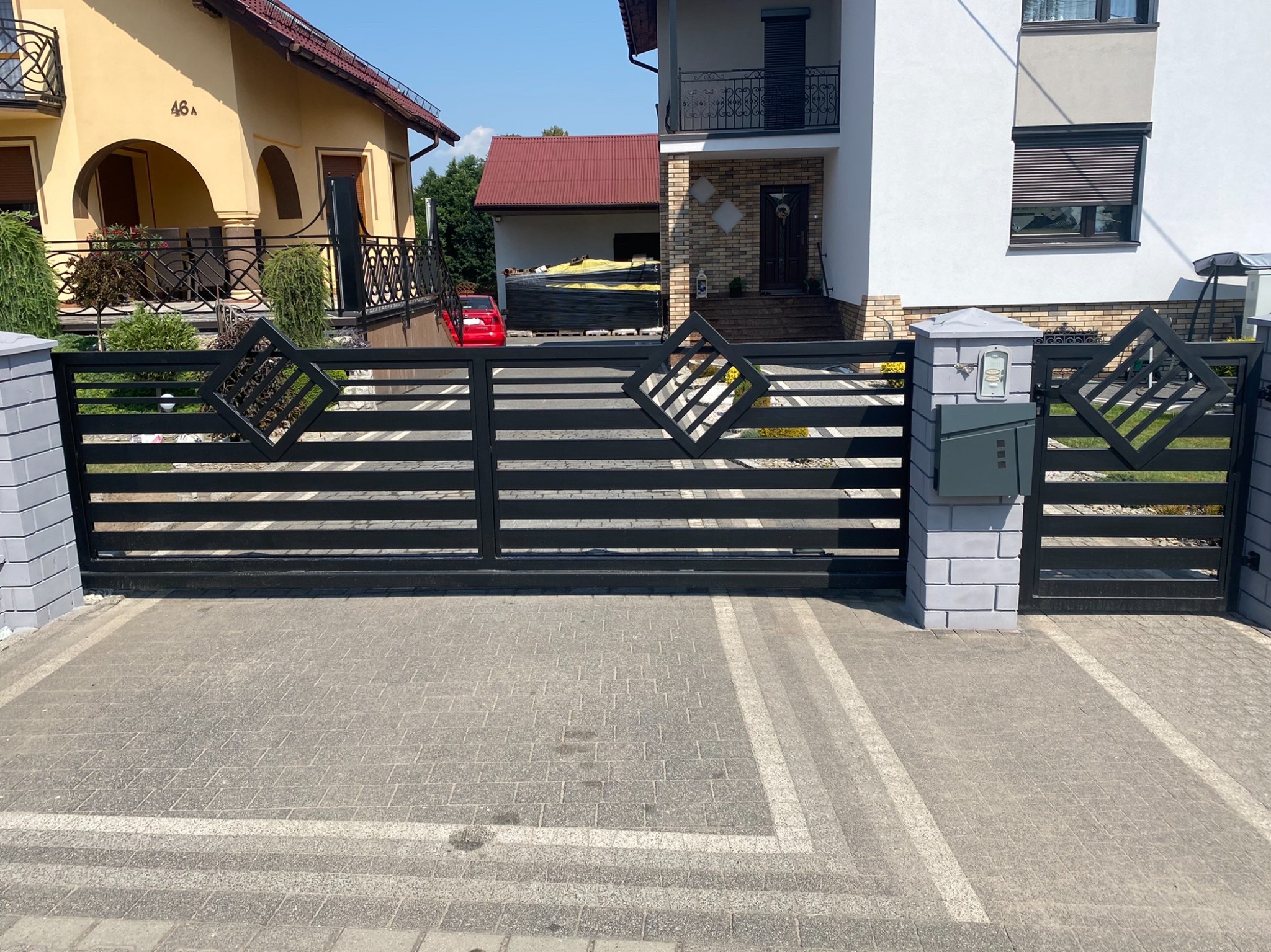 Czarna, pozioma brama wjazdowa z dekoracyjnymi rombami na tle dwóch domów z kostką brukową.