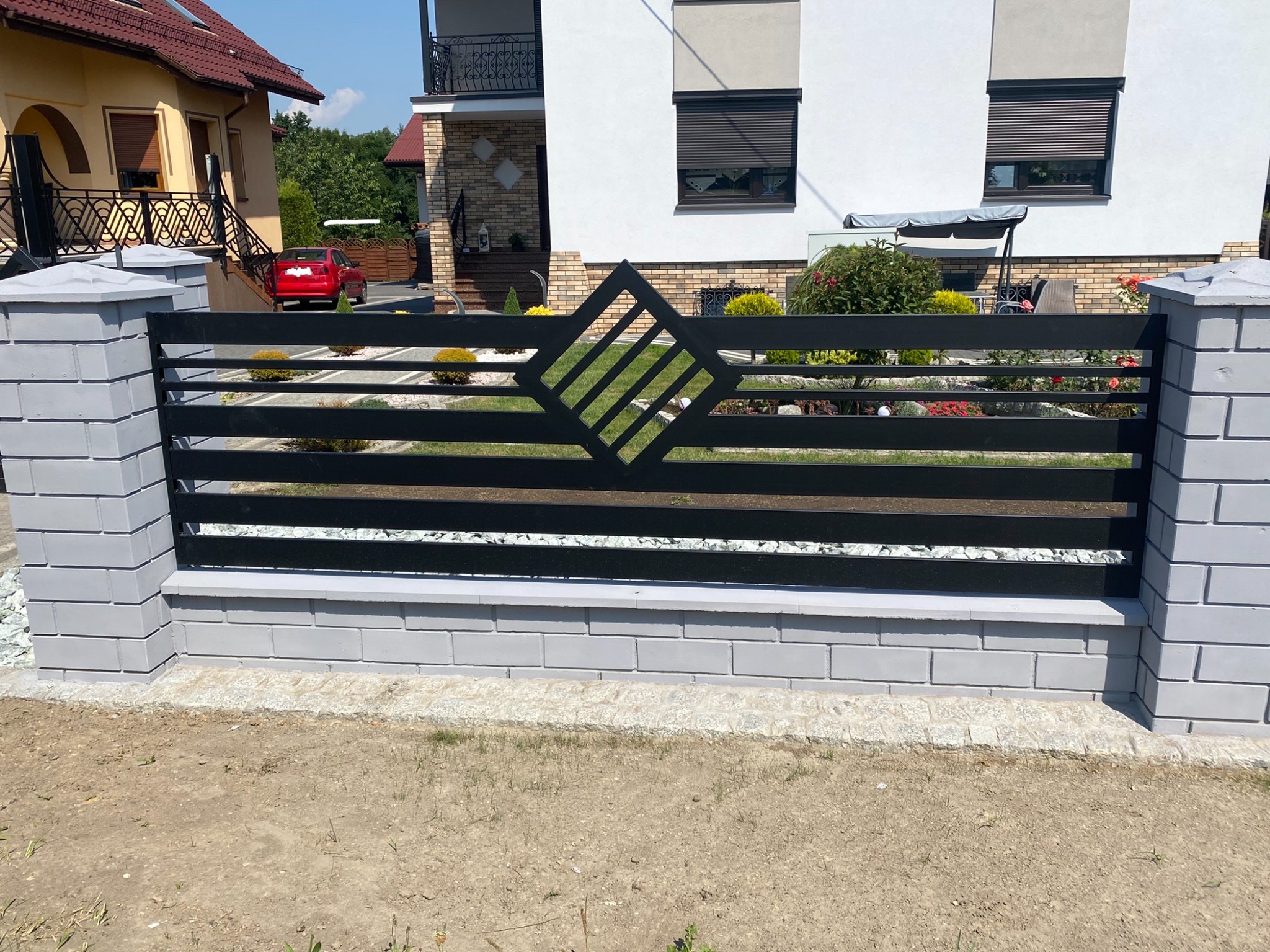 Nowoczesne, poziome przęsło z czarnych, metalowych profili z dekoracyjnym rombem, osadzone na szarym, murowanym cokole i słupkach, na tle domu z ogrodem.