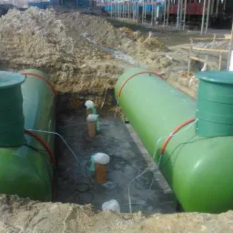 Dwa zielone zbiorniki zakopane w ziemi, prawdopodobnie na gaz płynny, z widocznymi włazami i przewodami, w wykopie z ziemią i konstrukcją w tle.
