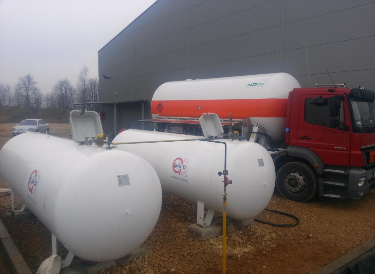 Białe zbiorniki na gaz z czerwonym logo producenta, jeden zbiornik załadowany na ciężarówkę z pomarańczowym pasem, widok na szary budynek w tle.