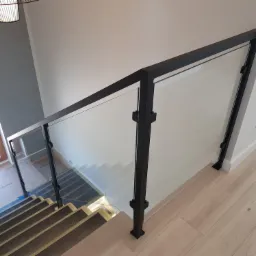 Szklana balustrada z czarnymi, metalowymi słupkami zabezpieczająca schody, widok z góry, częściowo zabezpieczone kartonem podczas remontu.