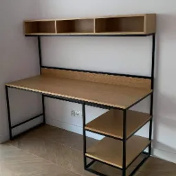 Nowoczesne biurko z blatem drewnianym i czarną metalową ramą, zintegrowane z półkami nad blatem i regałem bocznym z trzema półkami, ustawione na parkiecie w jodełkę.