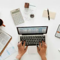 Widok z góry na biurko z laptopem, na którym widoczny jest arkusz kalkulacyjny, dłonie piszące na klawiaturze, gazeta biznesowa, telefon z aplikacją giełdową, kalkulator, notatnik, długopis...