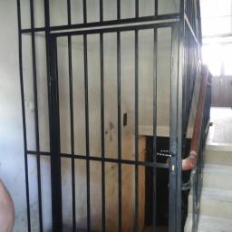 Fabro-met - Metalowa krata zabezpieczająca wejście na klatkę schodową, pomalowana na czarno, z widoczną klamką i fragmentem schodów.