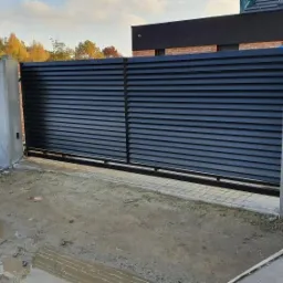 Nowoczesna, przesuwna brama wjazdowa z poziomych, czarnych lameli, zamontowana między dwoma betonowymi słupkami, z widocznymi przewodami elektrycznymi po lewej stronie i mokrym, ziemistym podłożem.