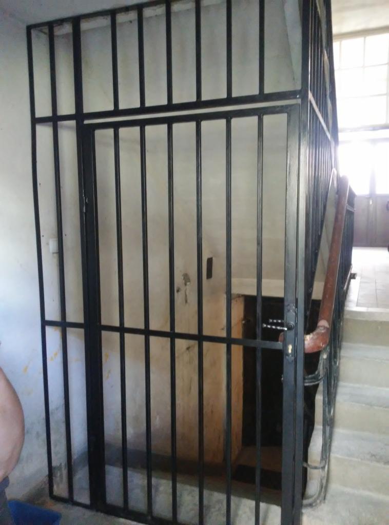 Metalowa krata zabezpieczająca wejście na klatkę schodową, pomalowana na czarno, z widoczną klamką i fragmentem schodów.