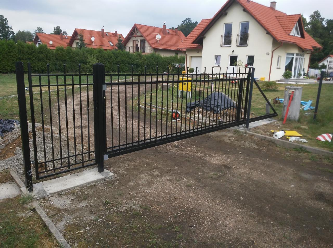 Czarna, metalowa brama wjazdowa z automatycznym napędem w trakcie montażu na tle domów jednorodzinnych z czerwonymi dachami.