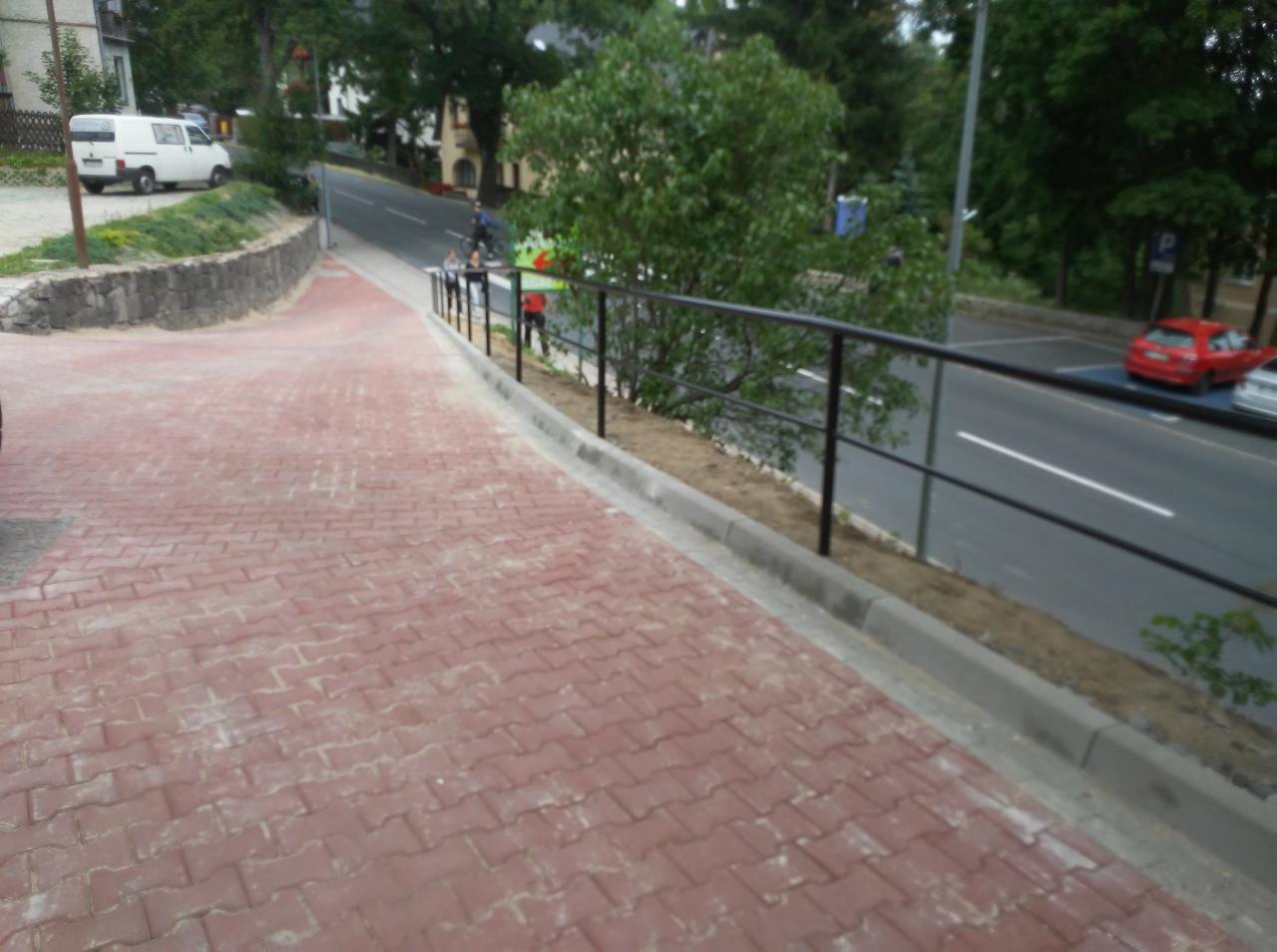 Czerwona kostka brukowa na pochyłym chodniku z nowo zamontowaną, czarną, metalową barierką ochronną wzdłuż ruchliwej ulicy.