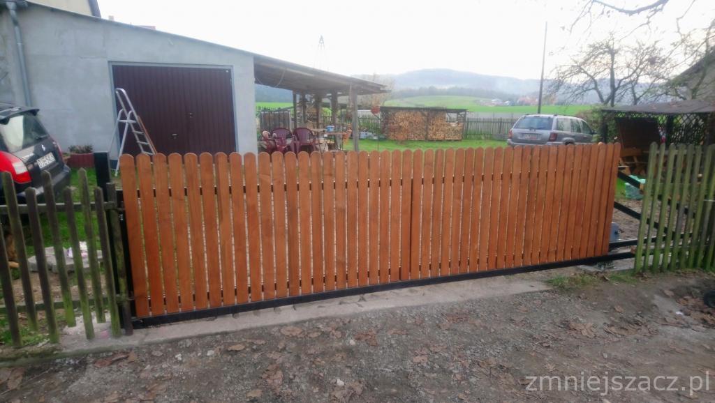 Nowa, drewniana brama wjazdowa z zaokrąglonymi sztachetami, osadzona na czarnej, metalowej ramie, widoczna na tle wiejskiego krajobrazu z budynkami gospodarczymi i samochodami.