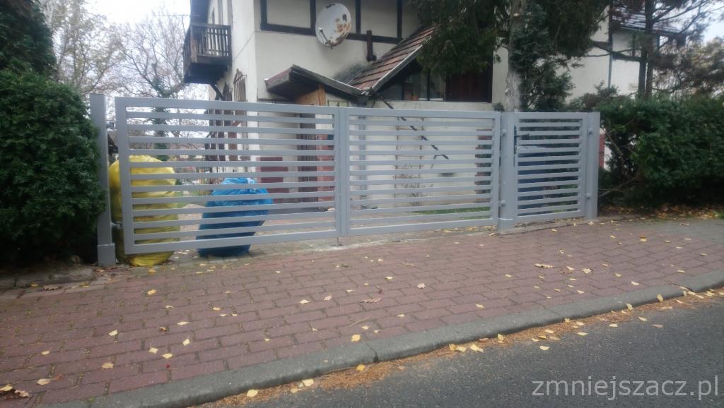 Nowoczesna brama wjazdowa z poziomych profili, pomalowana na szaro, z widocznymi workami na śmieci w kolorze żółtym i niebieskim po lewej stronie, na tle domu z balkonem i anteną satelitarną.