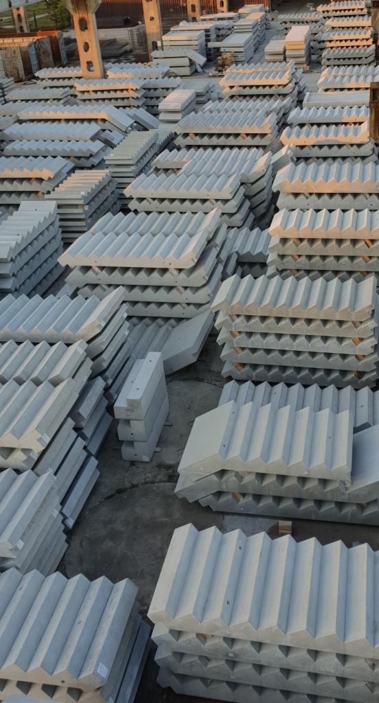 Stosy szarych, betonowych prefabrykatów schodowych o ząbkowanej strukturze, ułożone na placu budowy w regularnych rzędach, oświetlone popołudniowym słońcem.