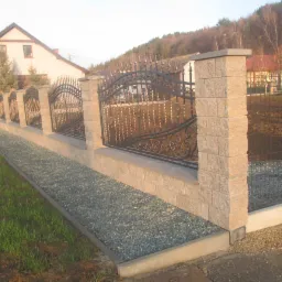 Ozdobny, czarny metalowy płot z kamiennymi słupkami oddziela posesję od trawnika i wysypanego kamieniami podłoża, w tle dom i zalesione wzgórze.