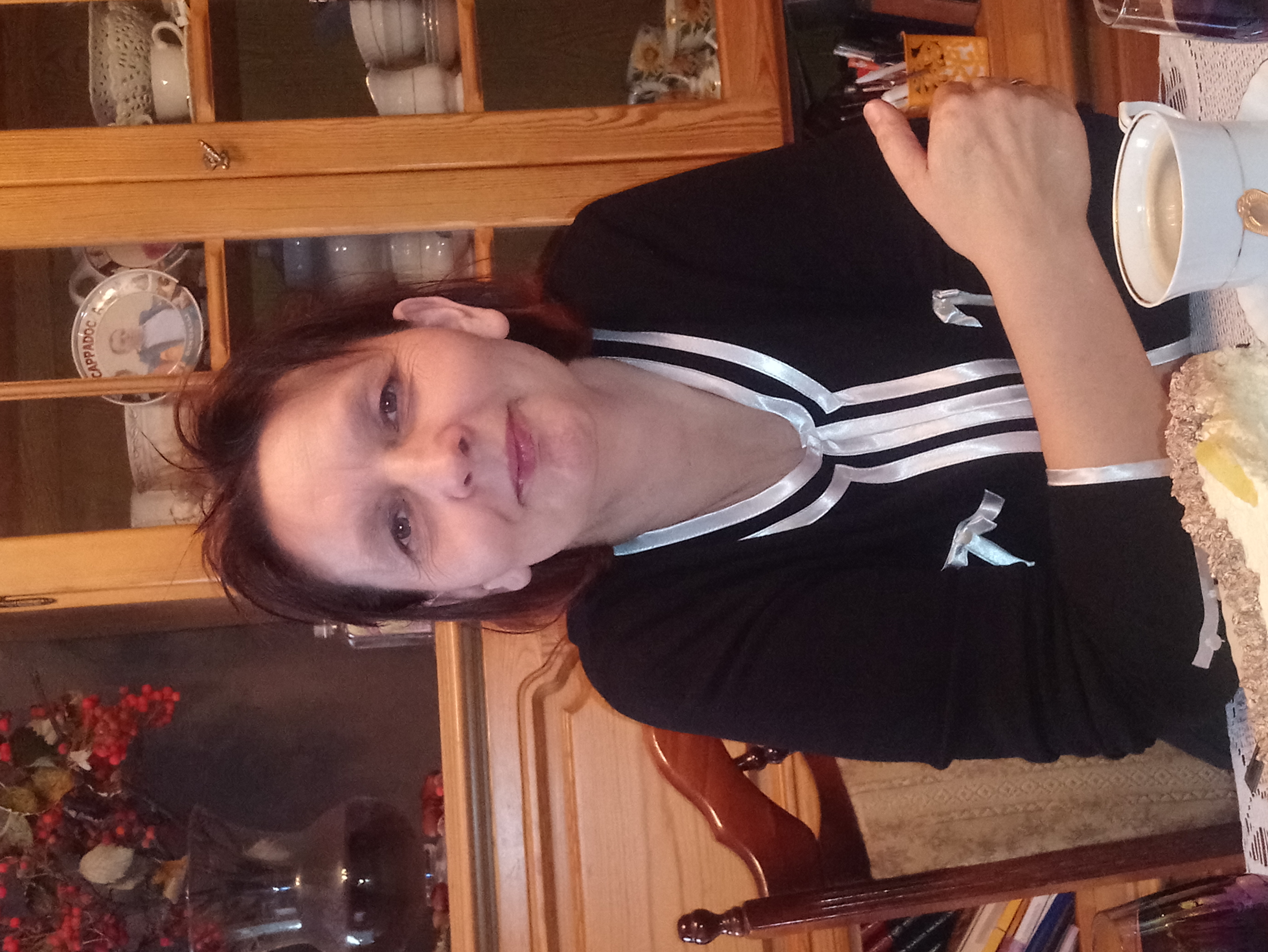 Kobieta w czarnej bluzce z białymi wstążkami siedzi przy stole z kawałkiem ciasta i filiżanką kawy, w tle drewniana szafka z ekspozycją filiżanek.