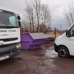 Dwa białe samochody dostawcze z logo firmy Planex parkują obok fioletowych kontenerów na gruz na błotnistym terenie w pochmurny dzień.