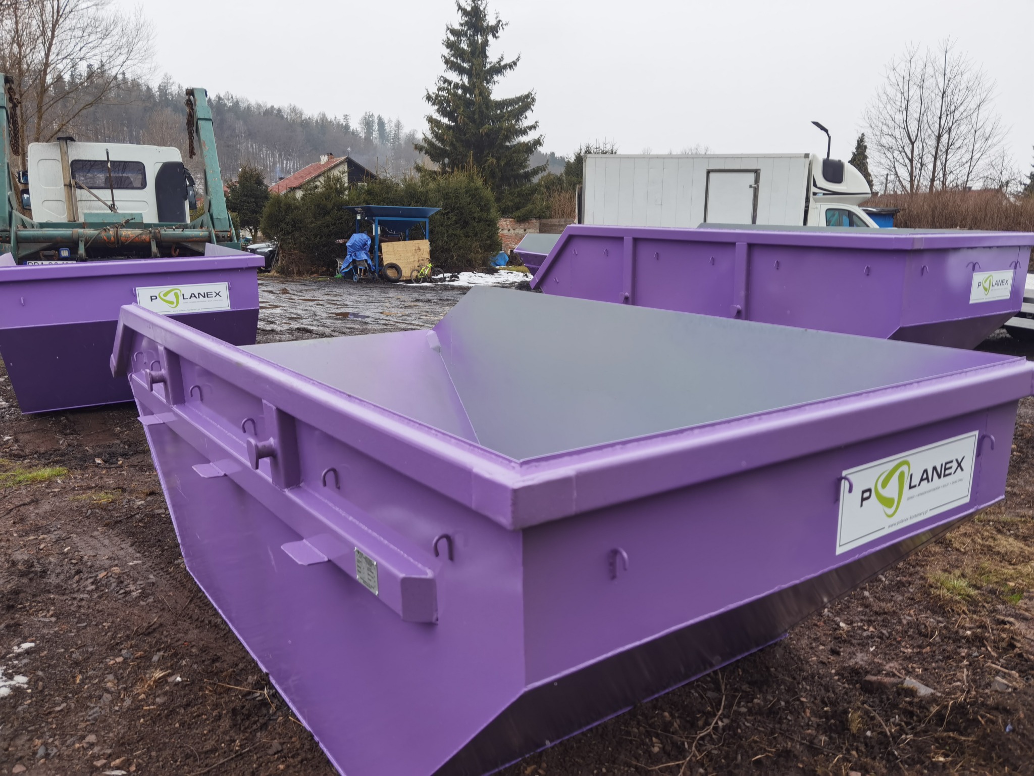 Fioletowe kontenery na gruz firmy Polanex, ustawione na błotnistym terenie w pochmurny dzień, z widocznym samochodem ciężarowym w tle.