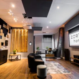 Nowoczesne biuro z drewnianą podłogą, ścianą z trójwymiarowych paneli w odcieniach beżu i czerni, ekranem projekcyjnym z napisem 'ZORTEA home' oraz minimalistycznym umeblowaniem, oświetlone...