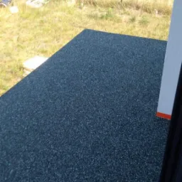 Ciemna, drobnoziarnista nawierzchnia balkonu z widokiem na zarośnięte pole i częściowo widoczne ogrodzenie.