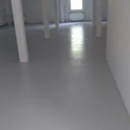 Jasnoszara, gładka podłoga w pustym, przemysłowym pomieszczeniu z białymi kolumnami i konstrukcją stropu. Okno w tle. Świeżo wylana i wypoziomowana powierzchnia.