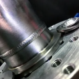 Precyzyjne spoiny na metalowym elemencie rurowym, widoczne ślady obróbki i połączenie śrubowe z podkładkami.