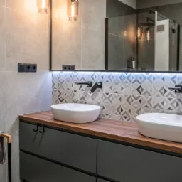 Nowoczesna łazienka z dwoma owalnymi umywalkami nablatowymi, czarnymi bateriami ściennymi, drewnianym blatem i szafką w kolorze grafitowym, podświetlane lustro oraz geometryczne płytki na ścianie.