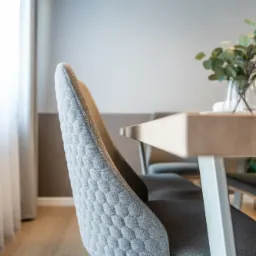 Szary, pikowany fotel przy drewnianym stole z bukietem eukaliptusa w szklanym wazonie, nowoczesne wnętrze jadalni z jasnym oknem.