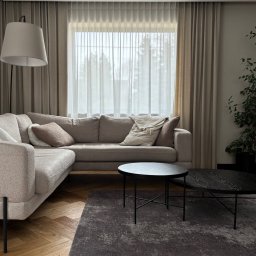Anna Kaczmarczyk Projektowanie Wnętrz - Nowoczesny salon z beżową narożną sofą, dwoma okrągłymi czarnymi stolikami kawowymi na ciemnym dywanie i drewnianą podłogą w jodełkę. Duże okno z beżowymi zasłonami i białą firaną oraz lampa...