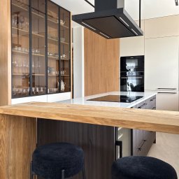Anna Kaczmarczyk Projektowanie Wnętrz - Nowoczesna kuchnia z wyspą kuchenną, czarnym okapem i płytą indukcyjną. Drewniany barek śniadaniowy z dwoma czarnymi hokerami. Szklane witryny z naczyniami i minimalistyczne oświetlenie liniowe.