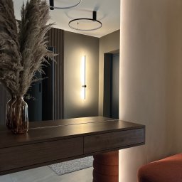 Anna Kaczmarczyk Projektowanie Wnętrz - Nowoczesny przedpokój w Krakowie z dużym lustrem, podwieszaną konsolą z ciemnego drewna, trawą pampasową i designerskim oświetleniem LED, tworzącym ciepłą atmosferę.