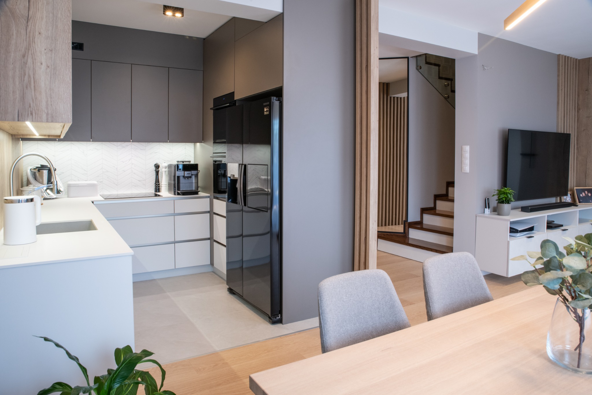Nowoczesna kuchnia z szarymi szafkami, białym blatem i czarną lodówką, widok na schody z drewnianymi stopniami i salon z telewizorem, stół z dwoma szarymi krzesłami i wazonem z zielonymi gałązkami...